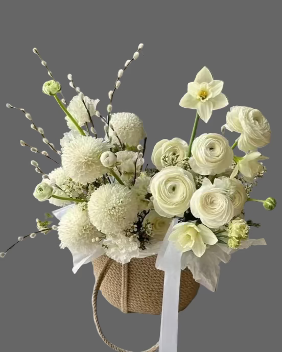 Elegant flower basket “Spring Whisper” featuring white ranunculus, daffodils, chrysanthemums, and seasonal blooms in a natural woven basket.