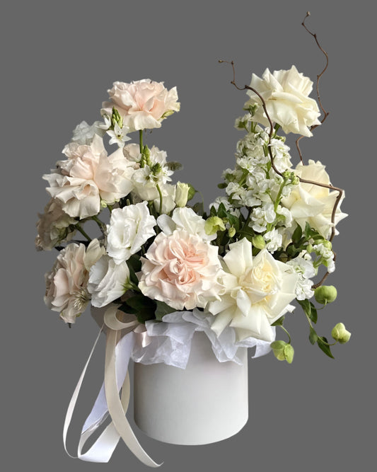 Elegant white and blush flower box with ivory roses, soft pink carnations, and seasonal greenery in a round hat box. Perfect floral gift for weddings, anniversaries, and birthdays. Available in multiple sizes.